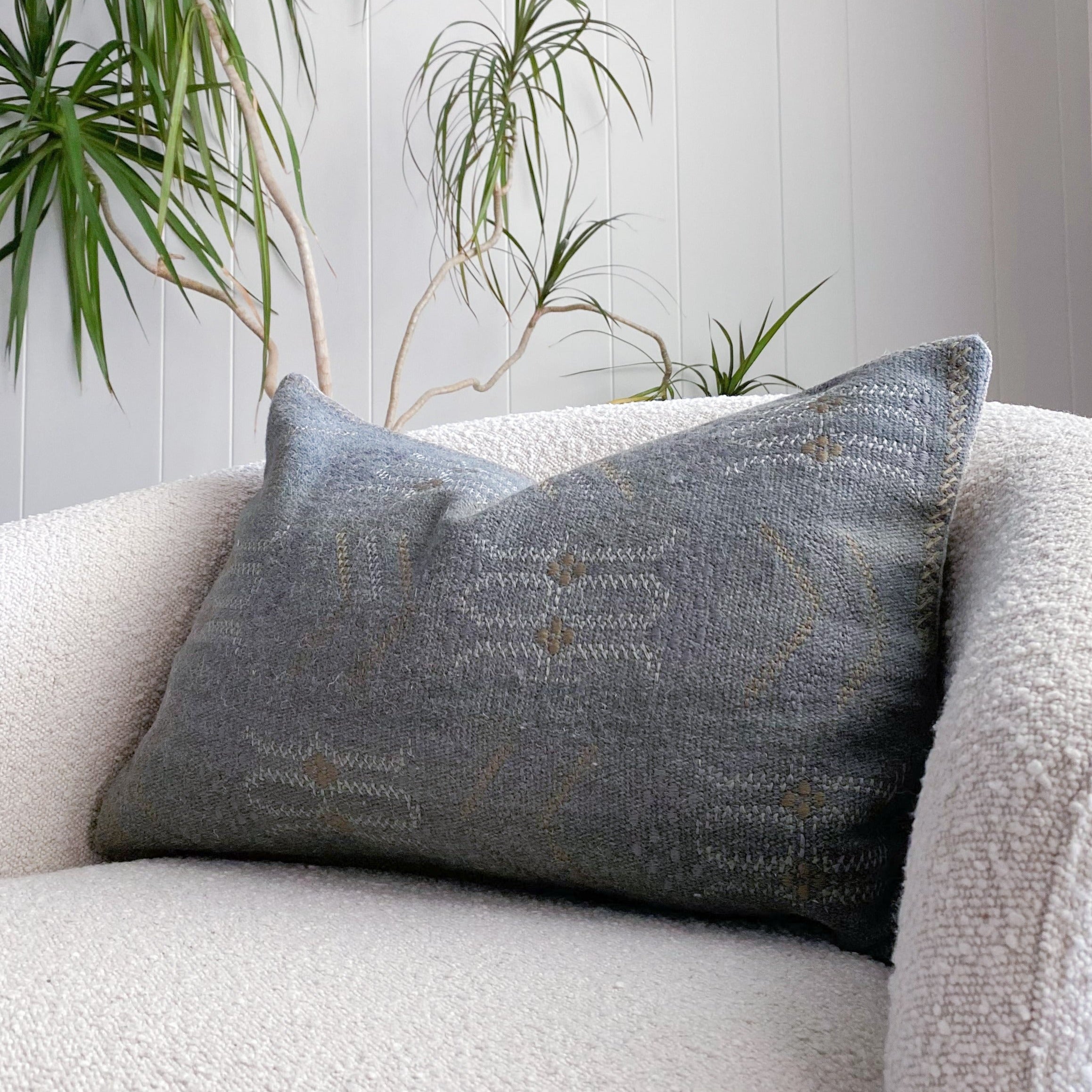 wool embroidered reversible lumbar throw pillow with a subtle dark teal, white and rust patterned front and a gray and mustard striped back. Shown sitting on an off-white boucle swivel arm chair in a white room with a decorative tree in the background